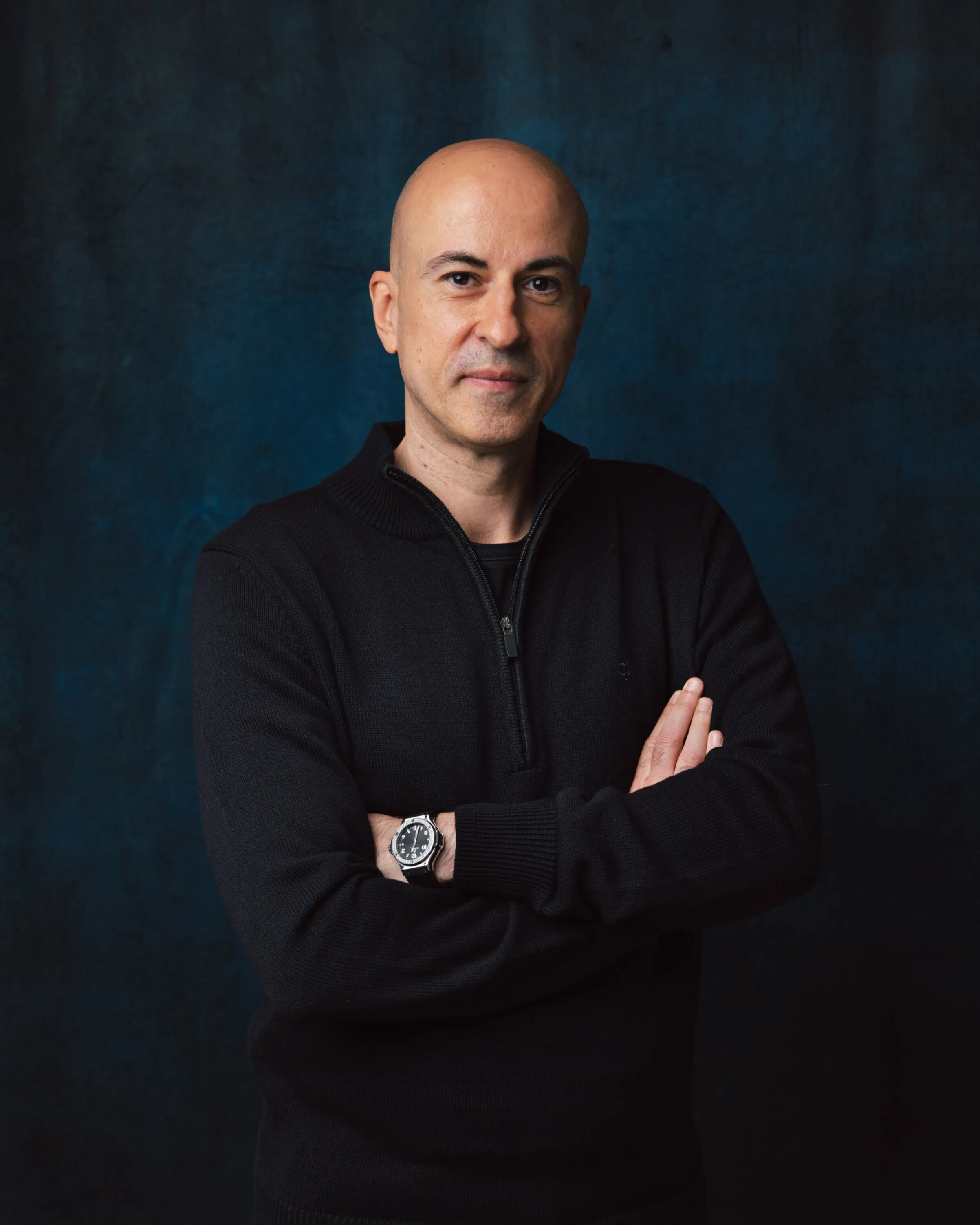 Corporate headshot of bald man in black zip jumper, arms crossed, dark background, London