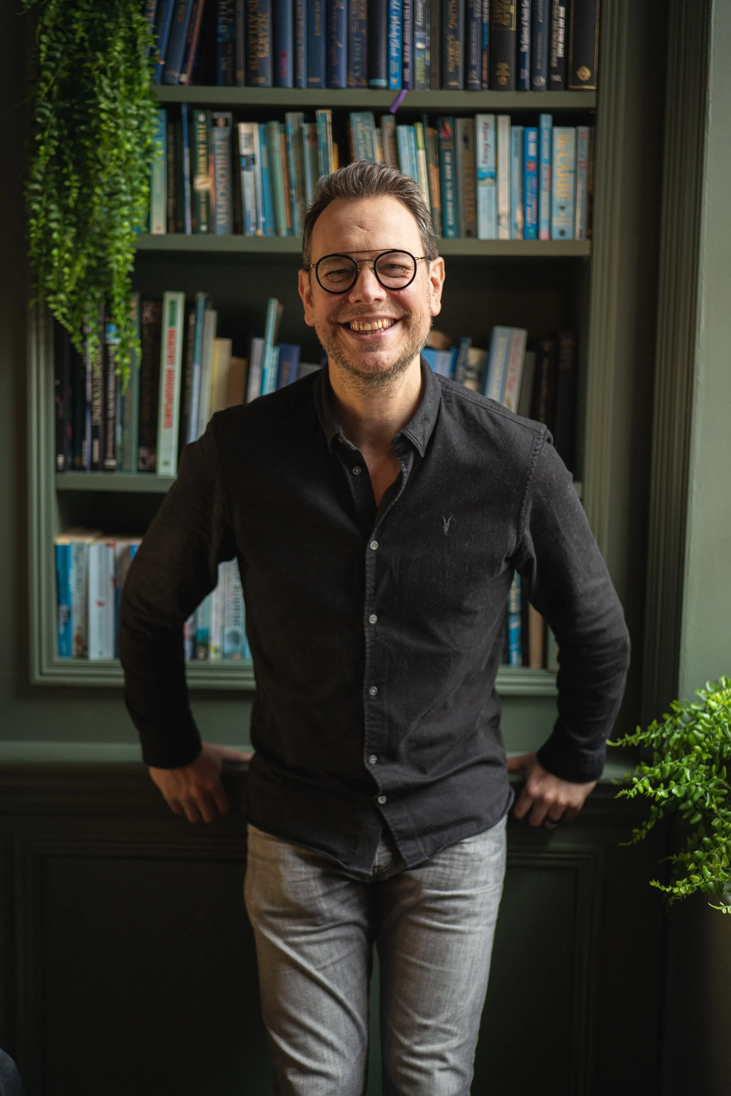 Corporate headshot of man in glasses and black shirt with bookshelf background, London